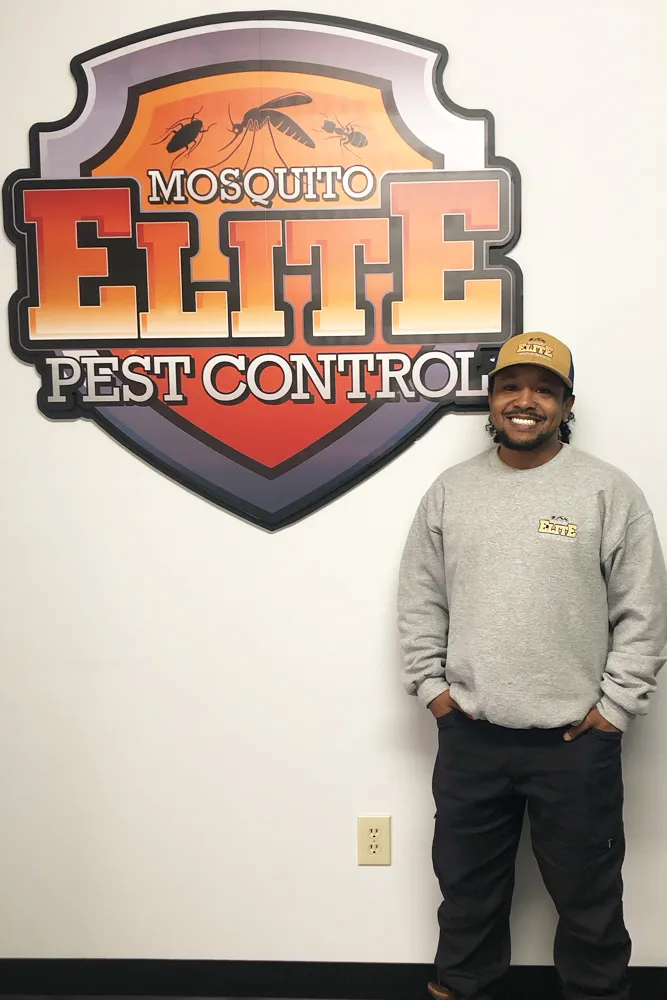 A smiling person standing in front of an "elite pest control" sign with logo, about to enter.